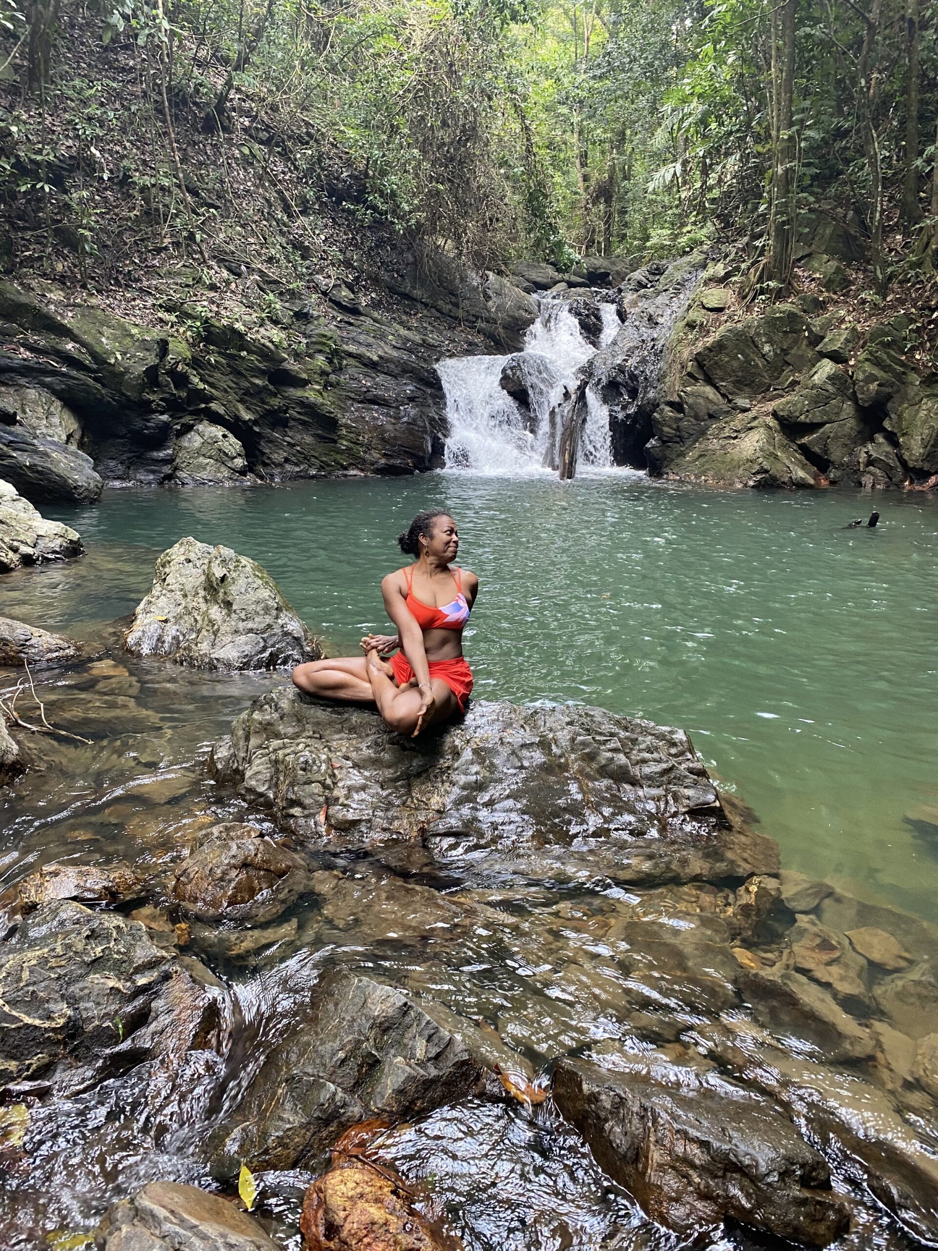 Penelope Kalloo sitting on rocks in a spinal twist yoga pose next to a waterfall