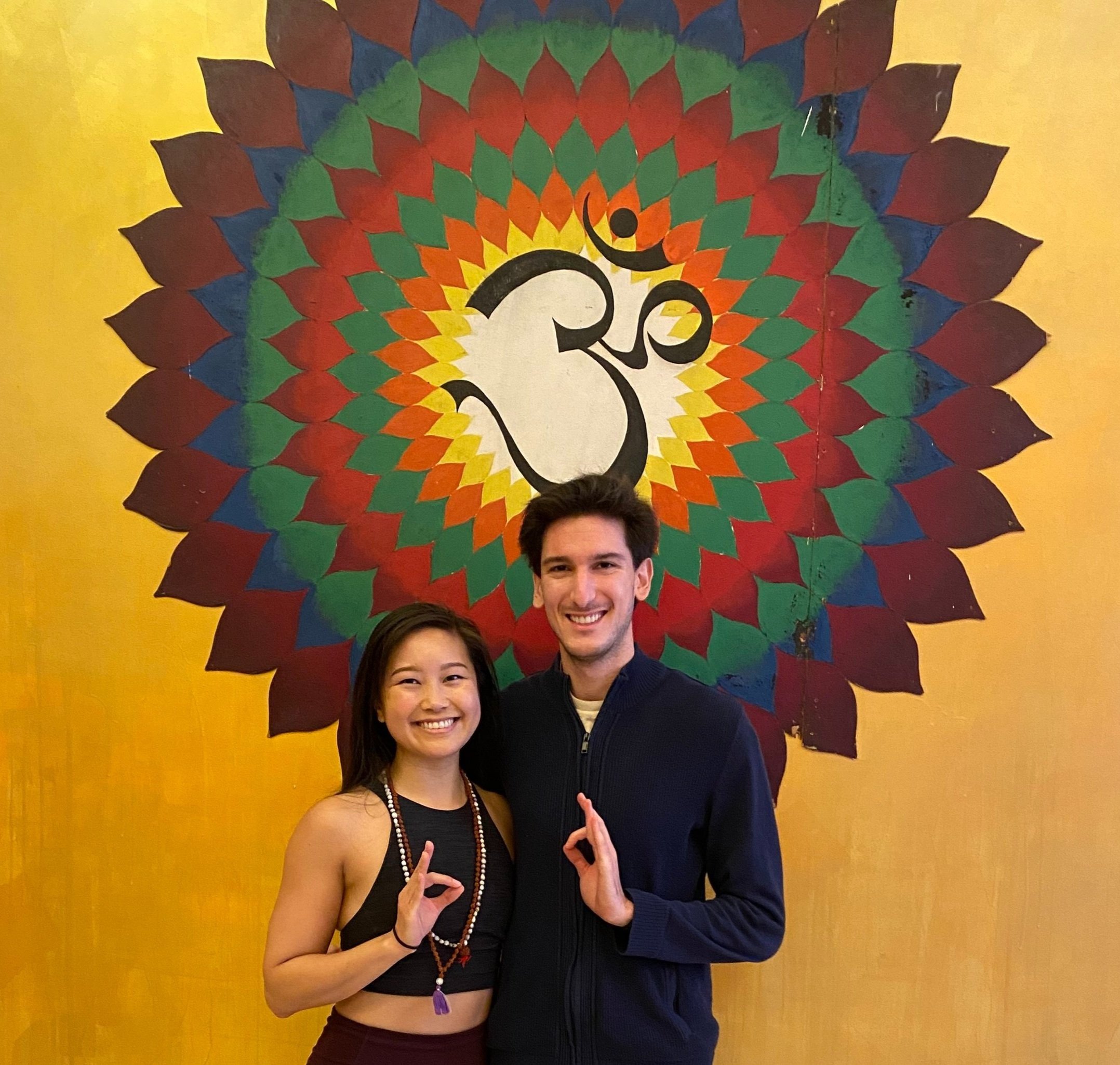 Yoga practitioners Dr. Jonathan Rosenthal and Jessica Hwang standing in front of an Om symbol, holding their hands in Gyana Mudra, a gesture of wisdom and inner peace.