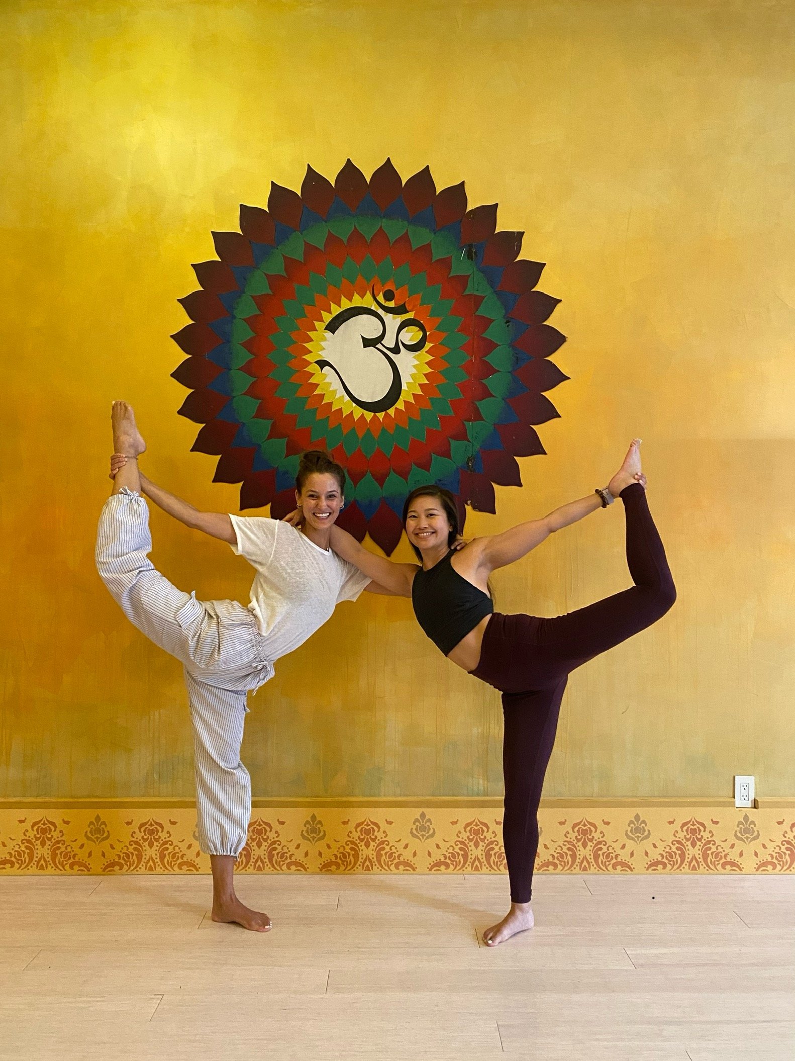 Victoria Davis and Jessica Hwang practicing standing bow yoga pose, with focused balance and grace, in front of a vibrant mural depicting the Om symbol.
