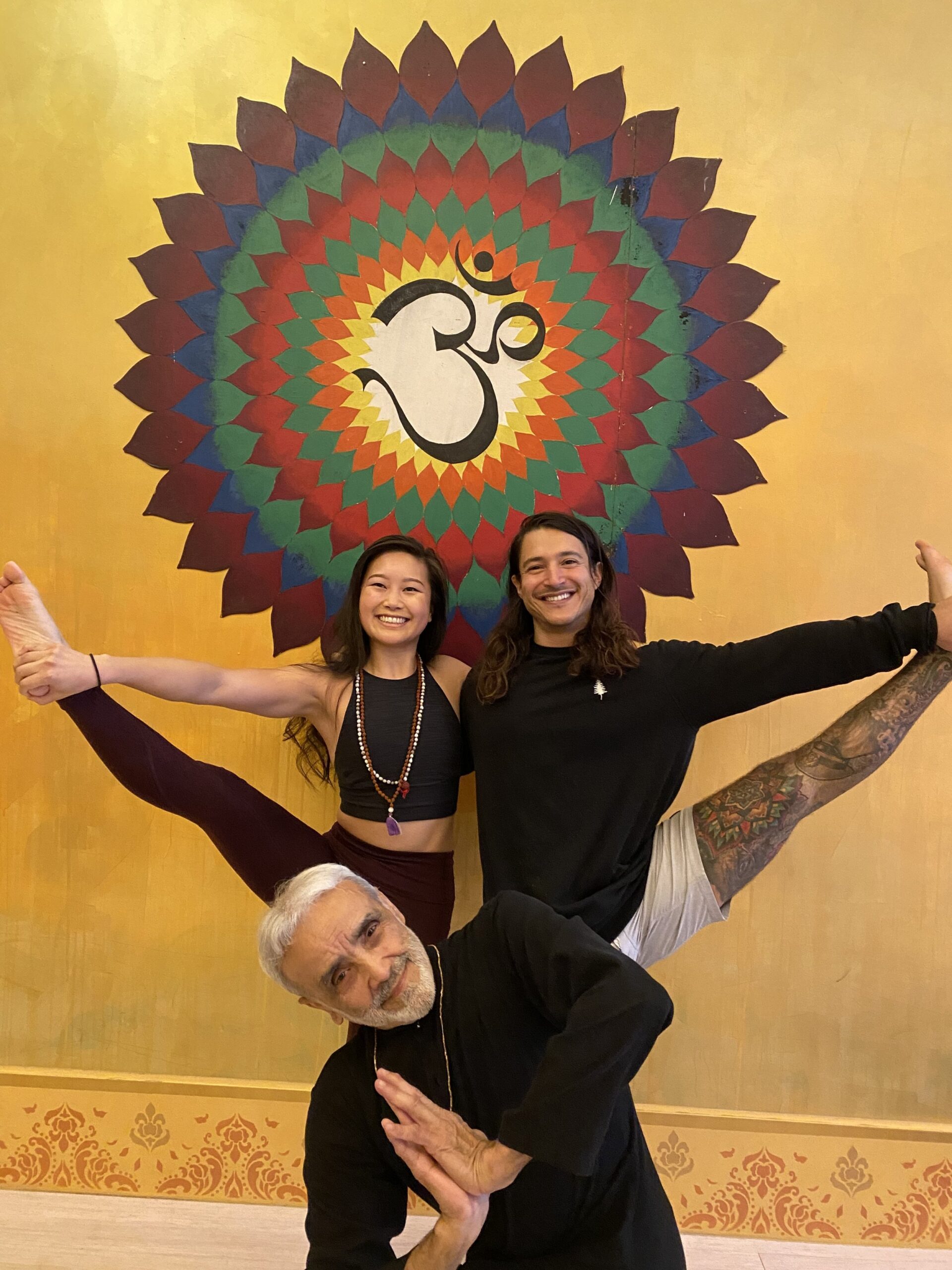 Yoga instructor Sri Dharma Mittra demonstrating a twist pose in front of Jessica Hwang and Rob Mendez holding ballet pose together, with a large Om symbol mural in the background of a serene yoga studio.
