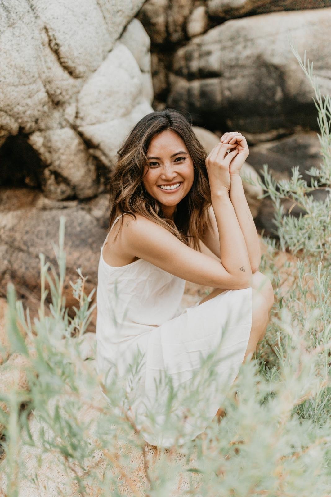 Lisa May Francisco crouching in lush green grass, looking thoughtfully at camera, embodying peaceful exploration in nature.