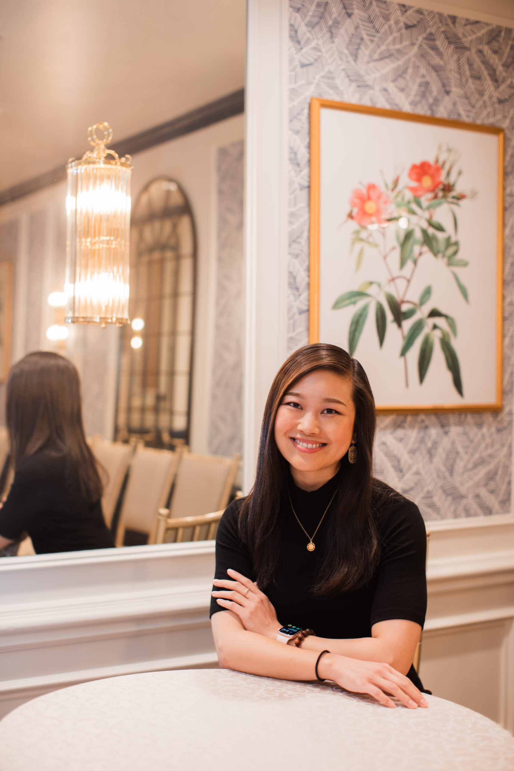 Jessica Hwang sitting at a table with arms folded in front of her and a mirror and flower painting behind her