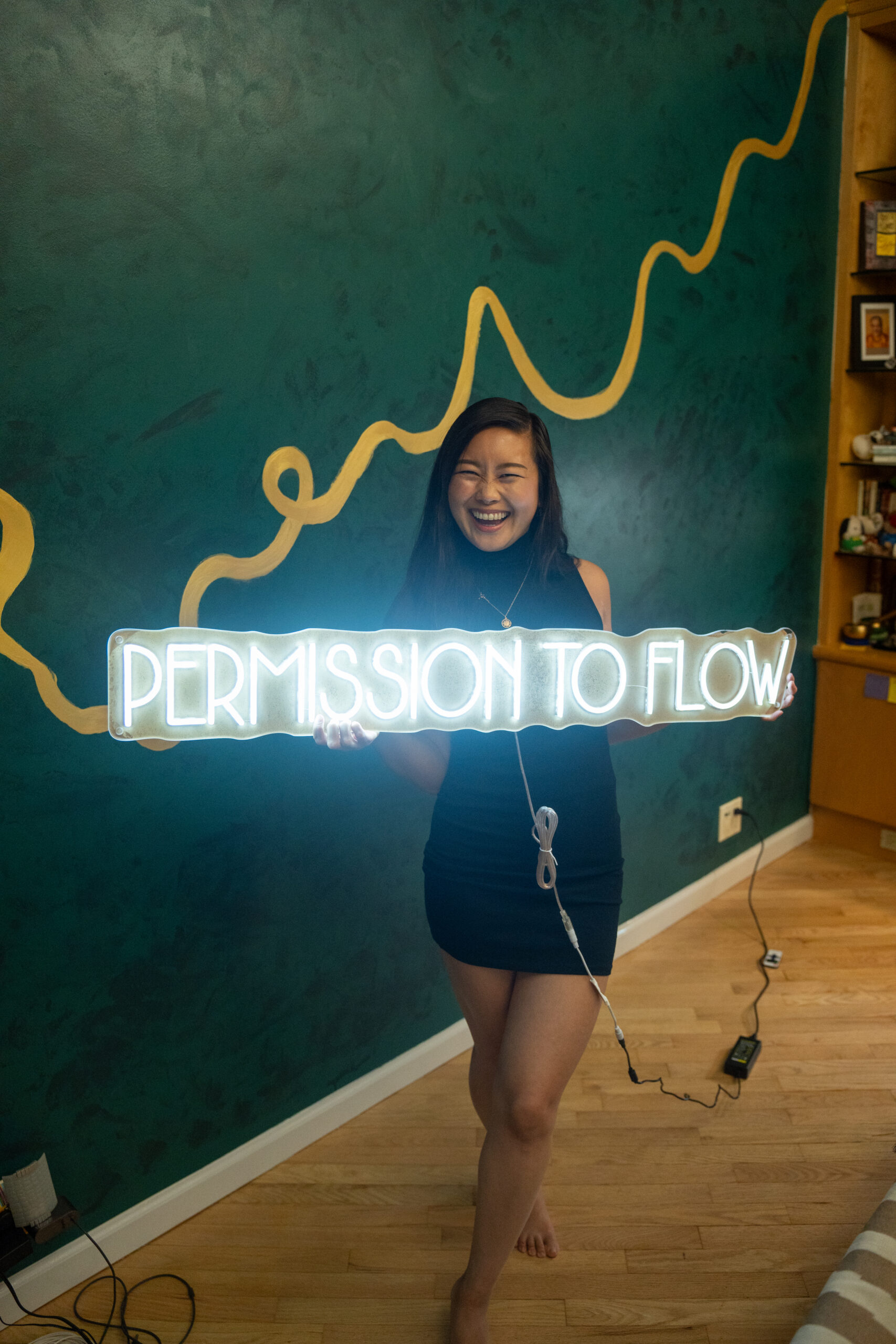 Jessica Hwang standing in front of her new wall mural holding up the neon sign for her podcast permission to flow